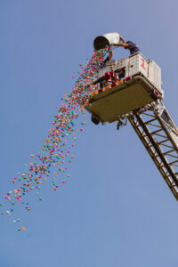 A firetruck's ladder is fully extended, high in the air, with only the clear, blue sky visible behind. Two firefighters, dressed in plain clothes, hold a large barrel and pour out 1000 small, colorful, rubber dolphins, which are showering down toward the camera.