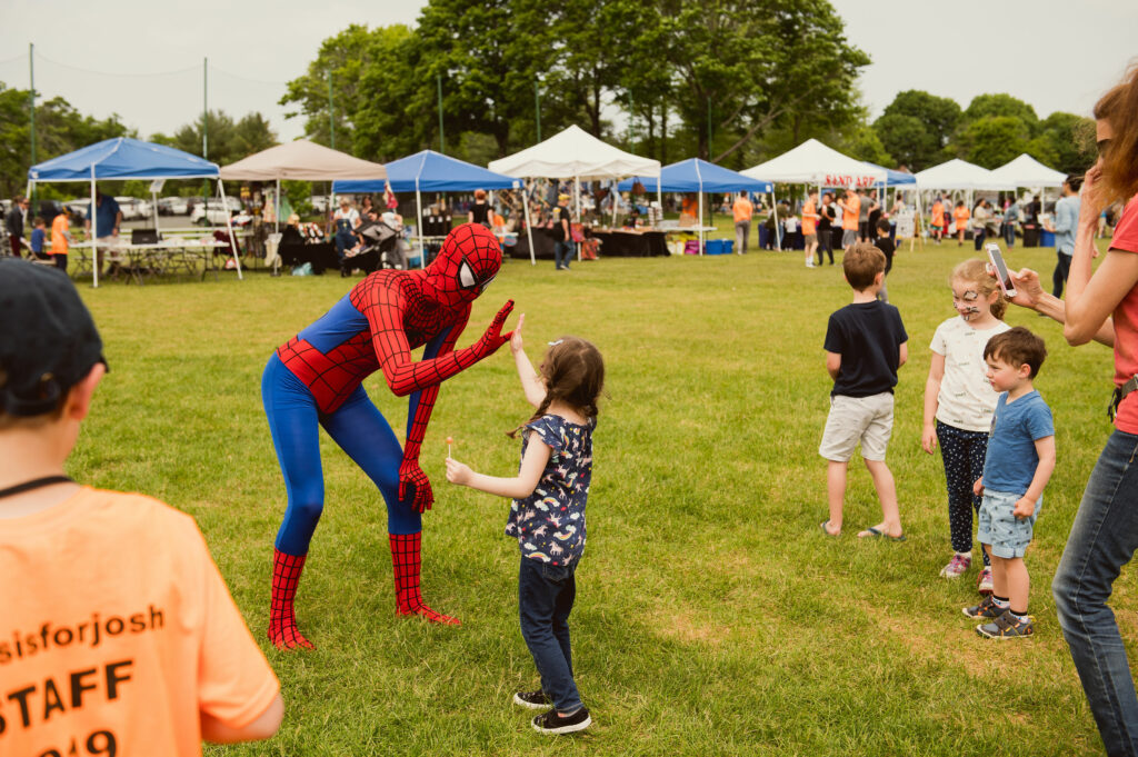 A small child is giving a high five to a person in a Spiderman costume. In the background, there are vendor tents at a festival. In the left corner, a boy is wearing a bright orange t-shirt that says "#ThisIsForJosh, Staff, 2019"